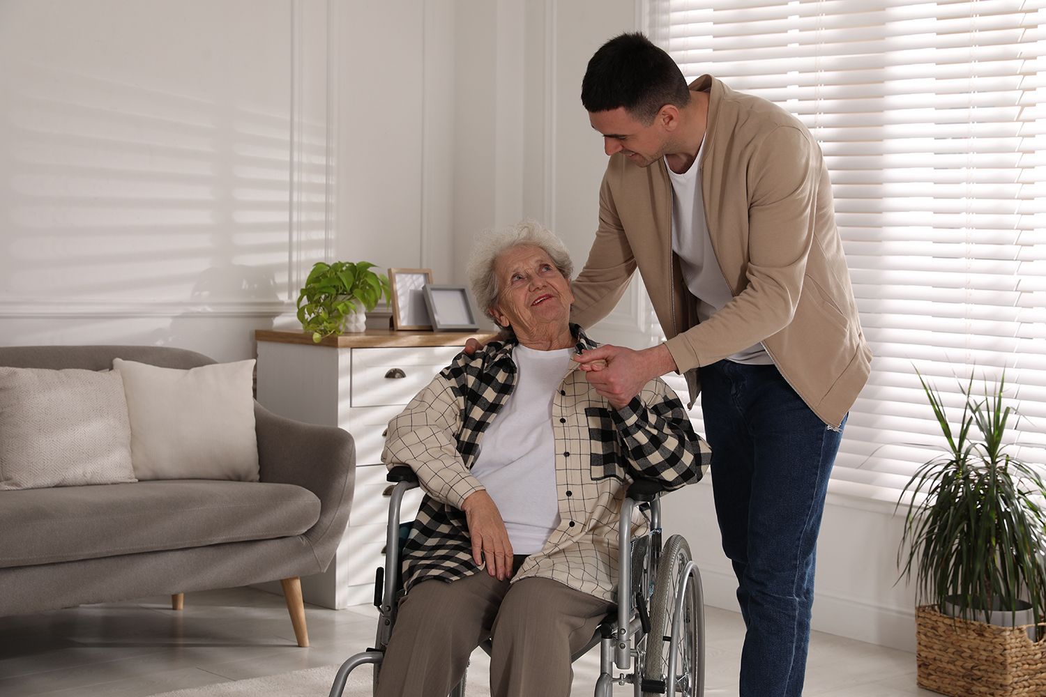 Man helping an elderly woman in a wheelchair in a bright living room