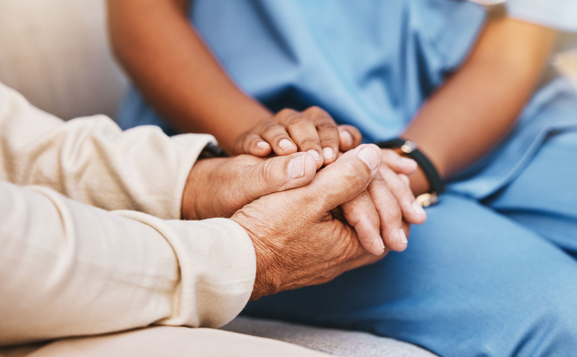 Hands clasped in comfort, with a caregiver in blue and an elderly person in beige sitting together.