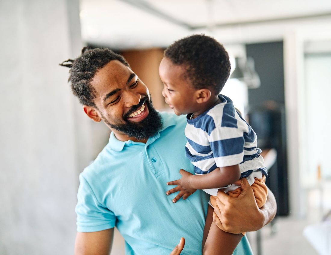 A smiling parent in a light blue polo shirt holds a small child who is looking at them in a brightly lit indoor space.