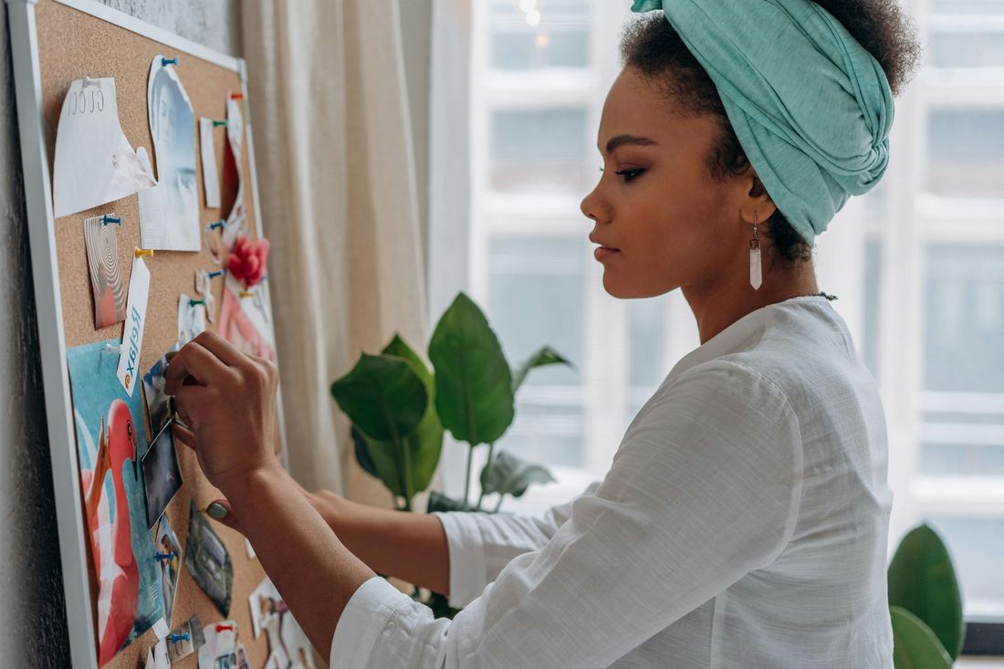 A person wearing a light blue headwrap and white shirt pins a picture onto a cork mood board in a brightly lit room.
