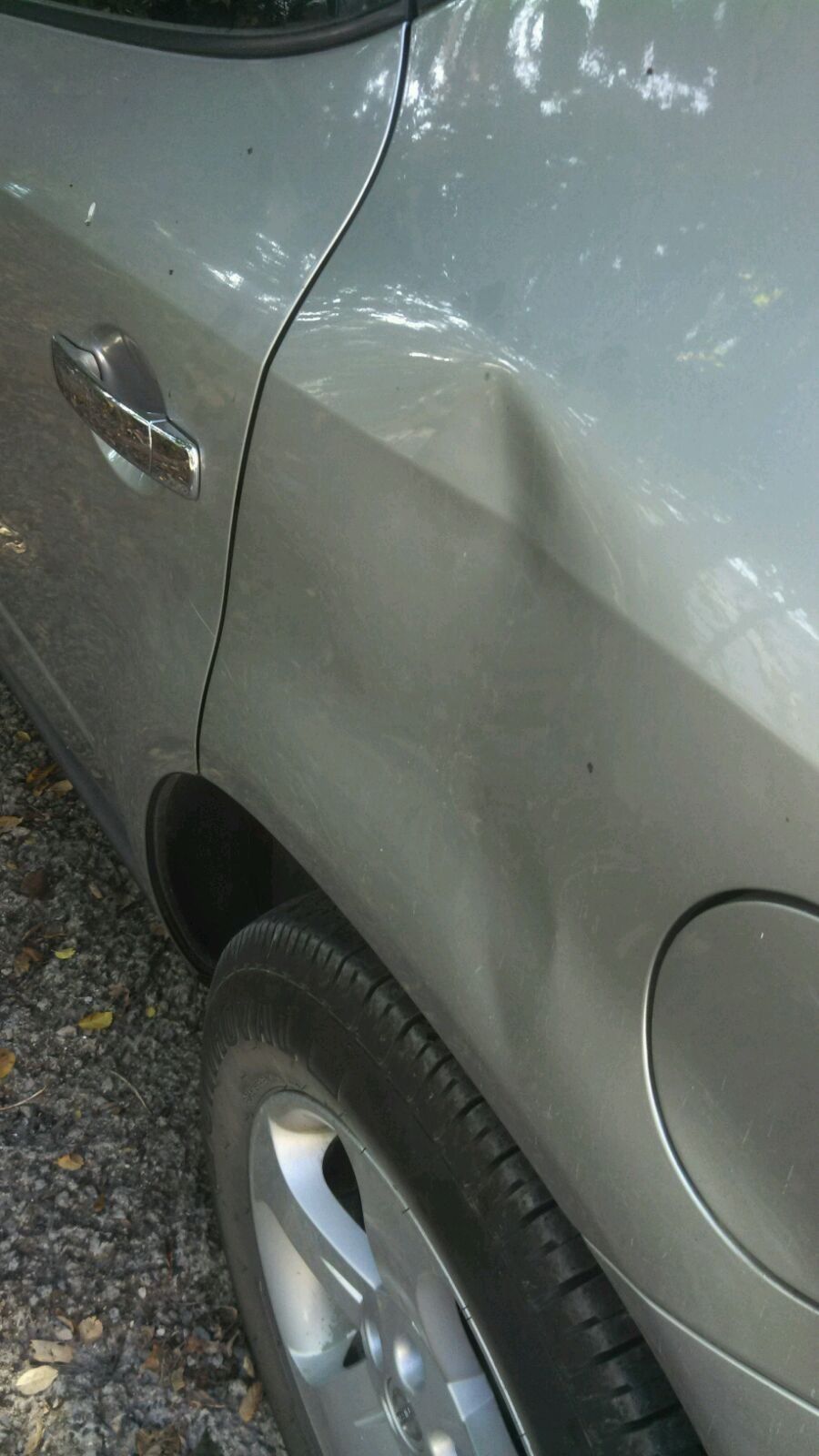A silver car with a damaged fender is parked on the side of the road.