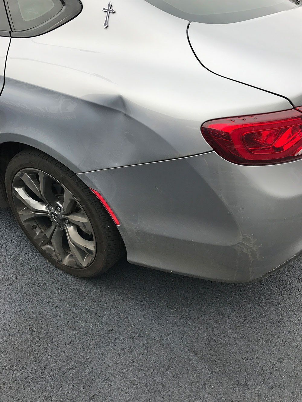 A silver car with a damaged bumper is parked on the side of the road.