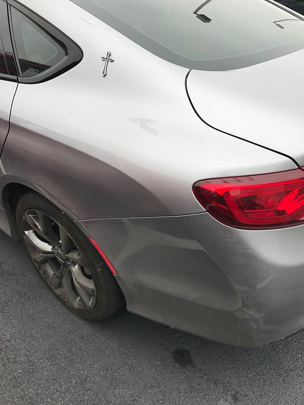 A silver car with a damaged bumper is parked on the side of the road.