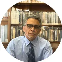 Man in glasses and blue shirt seated in front of bookshelves, looking at the camera.