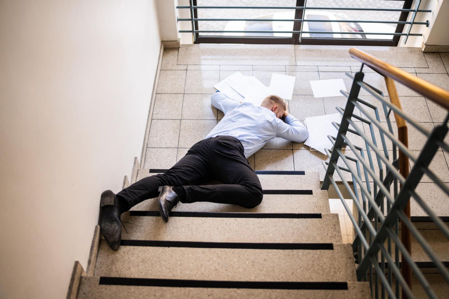 A person in a light blue shirt and dark trousers lies face-down on a flight of stairs surrounded by scattered papers.