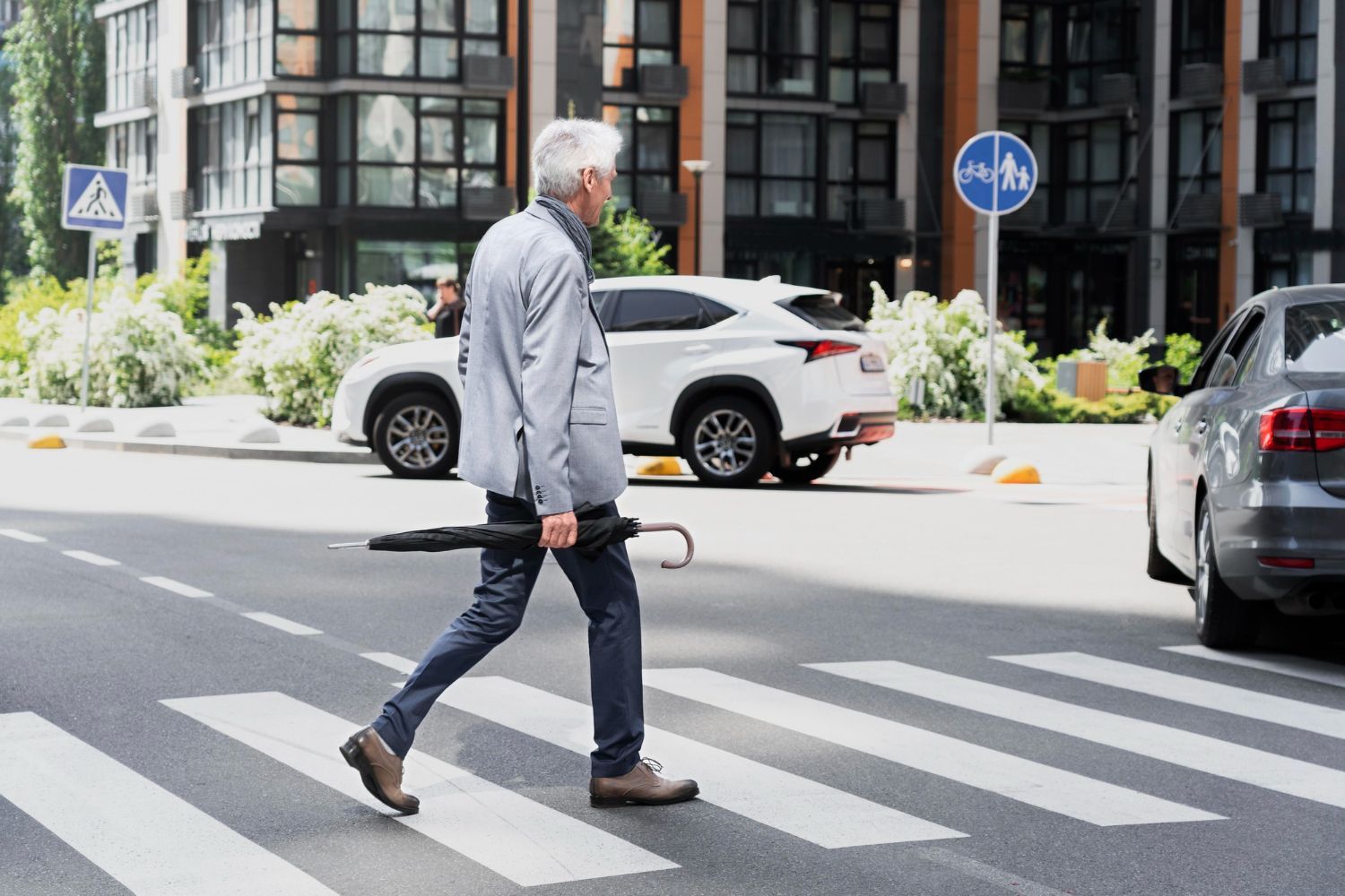 A person carrying a folded umbrella walks across a city street on a white-striped pedestrian crosswalk.