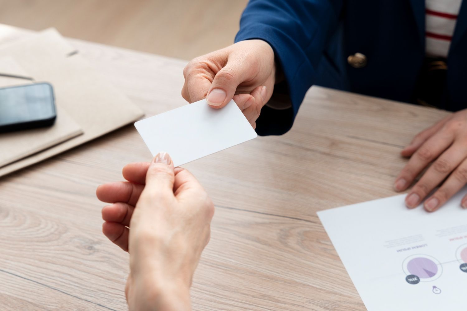 Two people sitting at a wooden desk exchange a blank white business card.