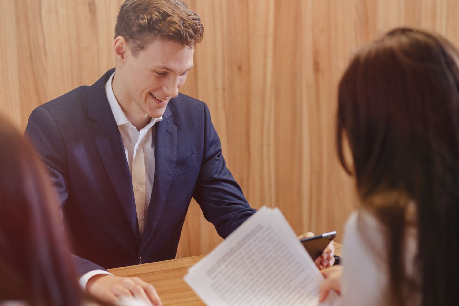 A man in a suit smiles during a meeting, looking at documents held by a person across a wooden table.