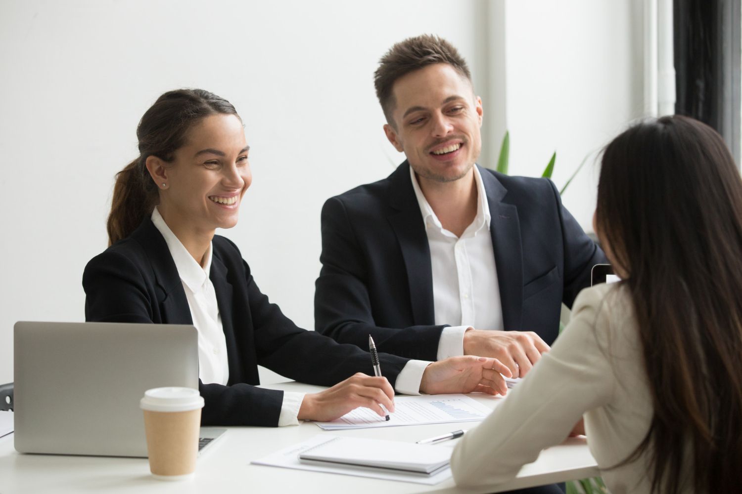 Two business professionals interview a candidate, all smiling while sitting at a table with a laptop and documents.