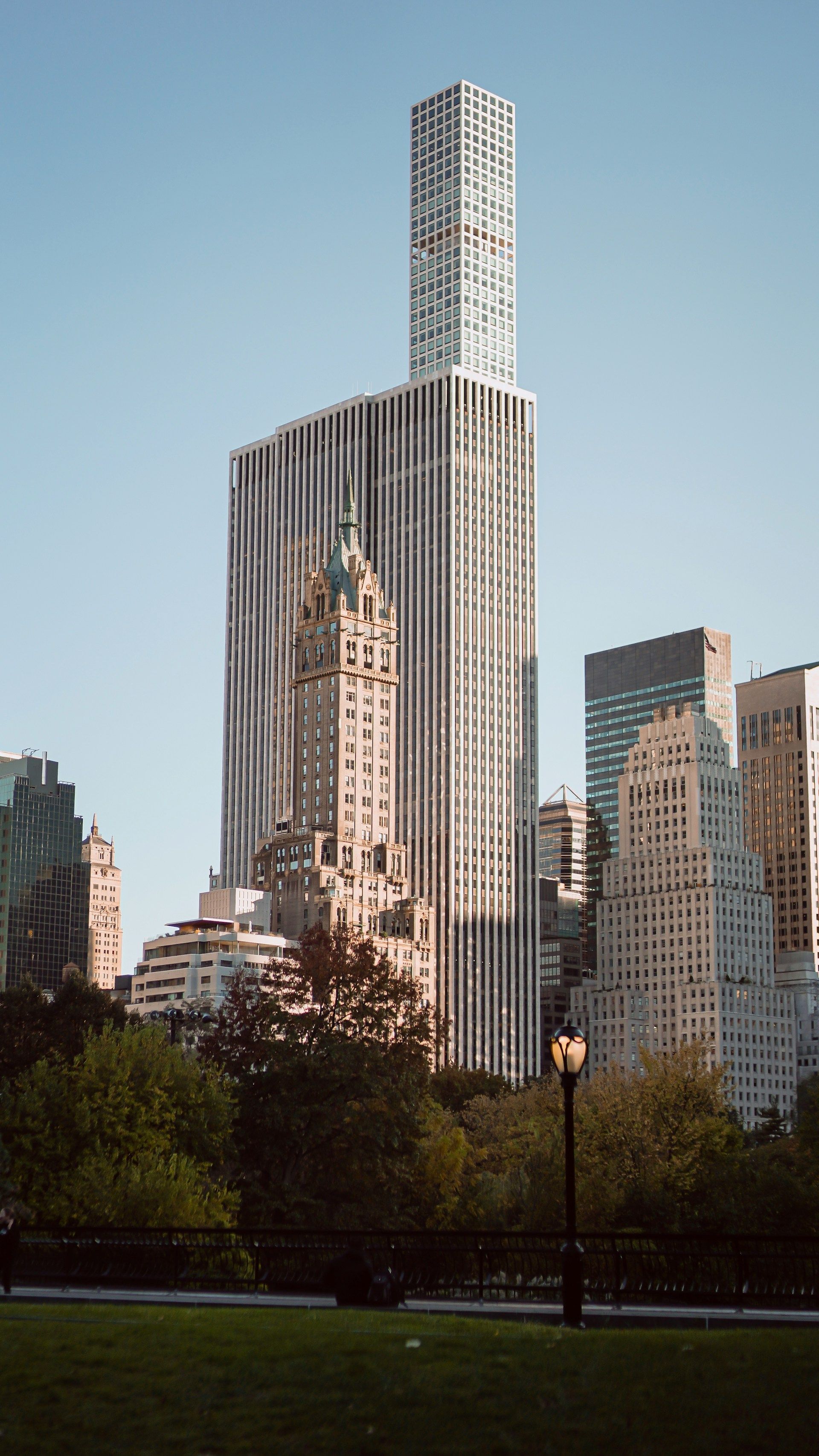 A tall, slender, modern skyscraper rises behind a historic ornate building, viewed from a park at dusk.
