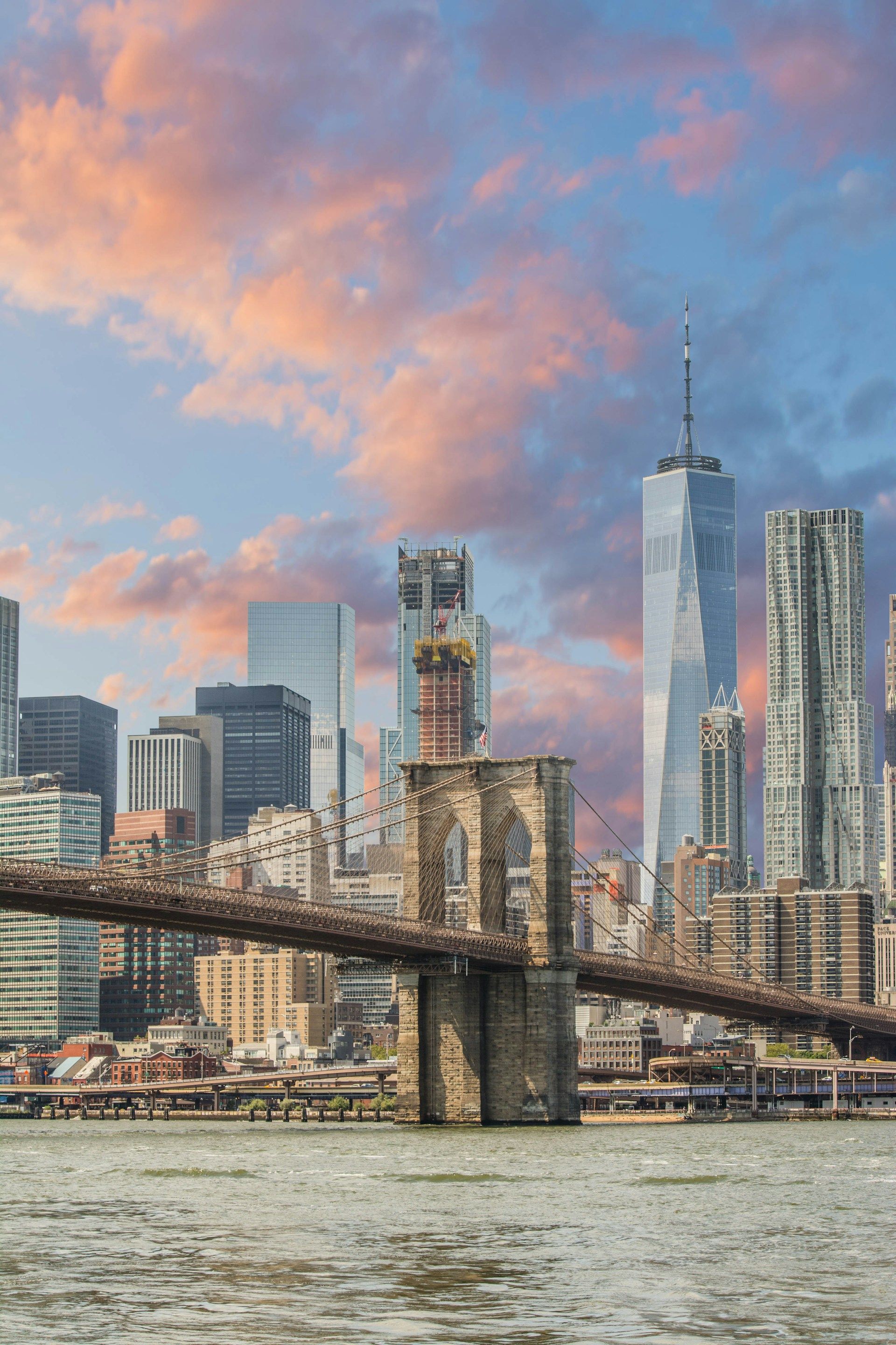 The Brooklyn Bridge spans across the foreground with the New York City skyline under a sunset sky in the background.