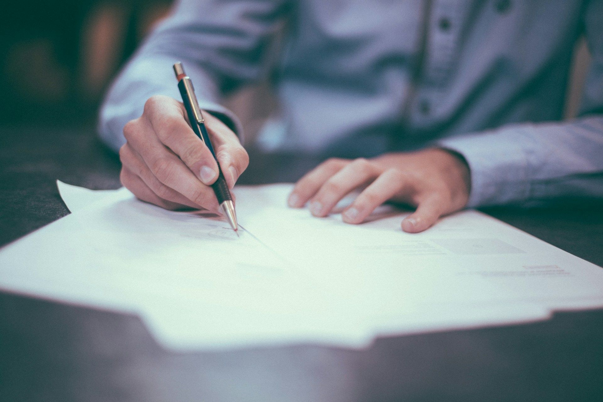 A person’s hands writing with a pen on a stack of white papers on a desk.