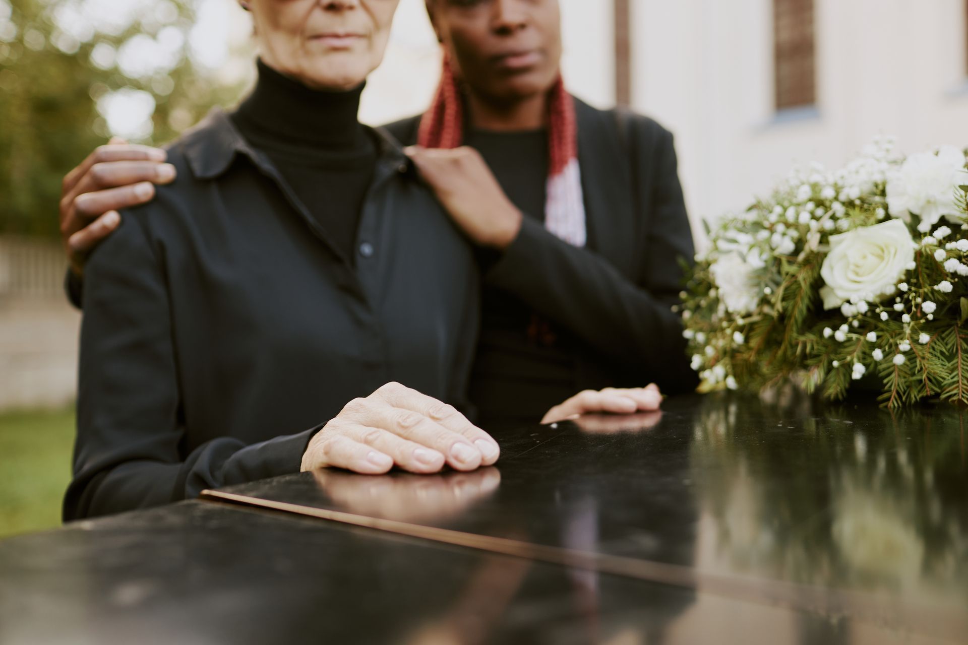 Two people in dark clothing standing by a coffin, with one placing a supportive hand on the other's shoulder.