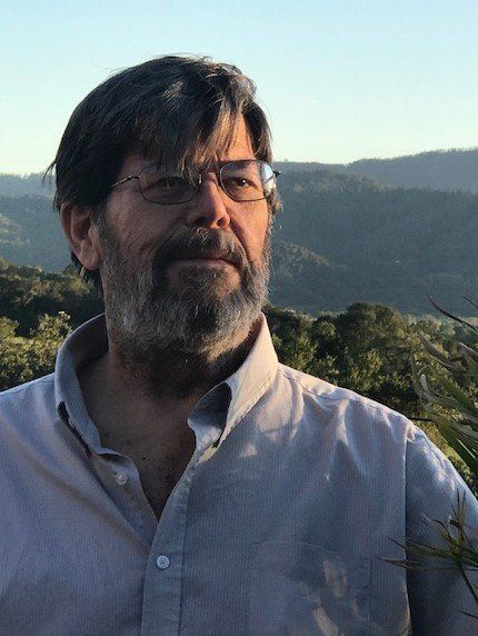 Man with beard and glasses, outdoors with a mountain backdrop.