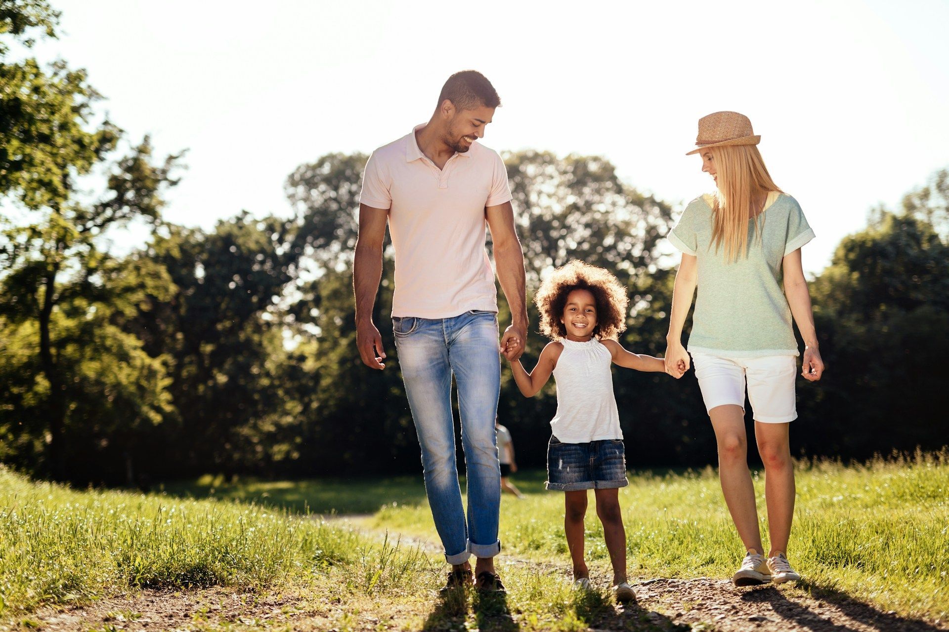 Family walking hand in hand through a sunny park, with a child between two adults.