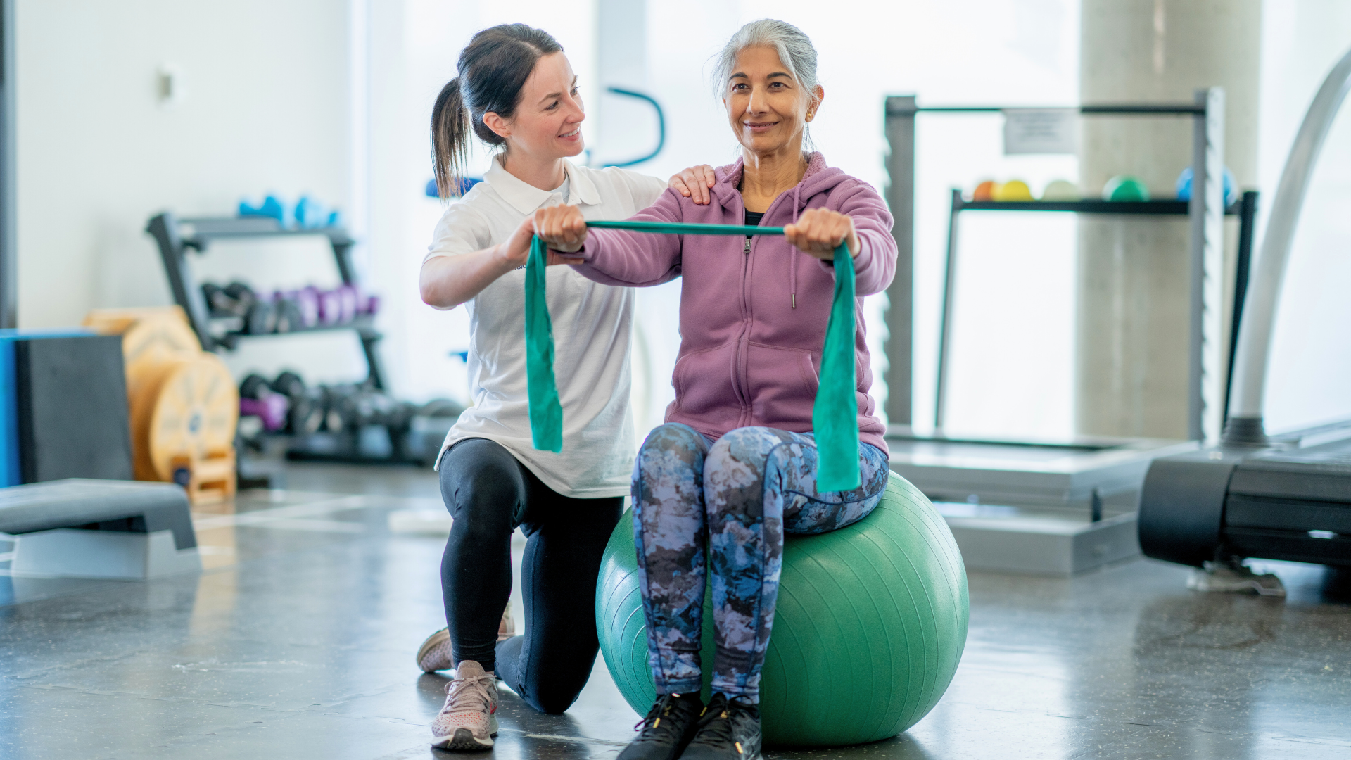 A woman is helping an older woman do exercises on an exercise ball in a gym.