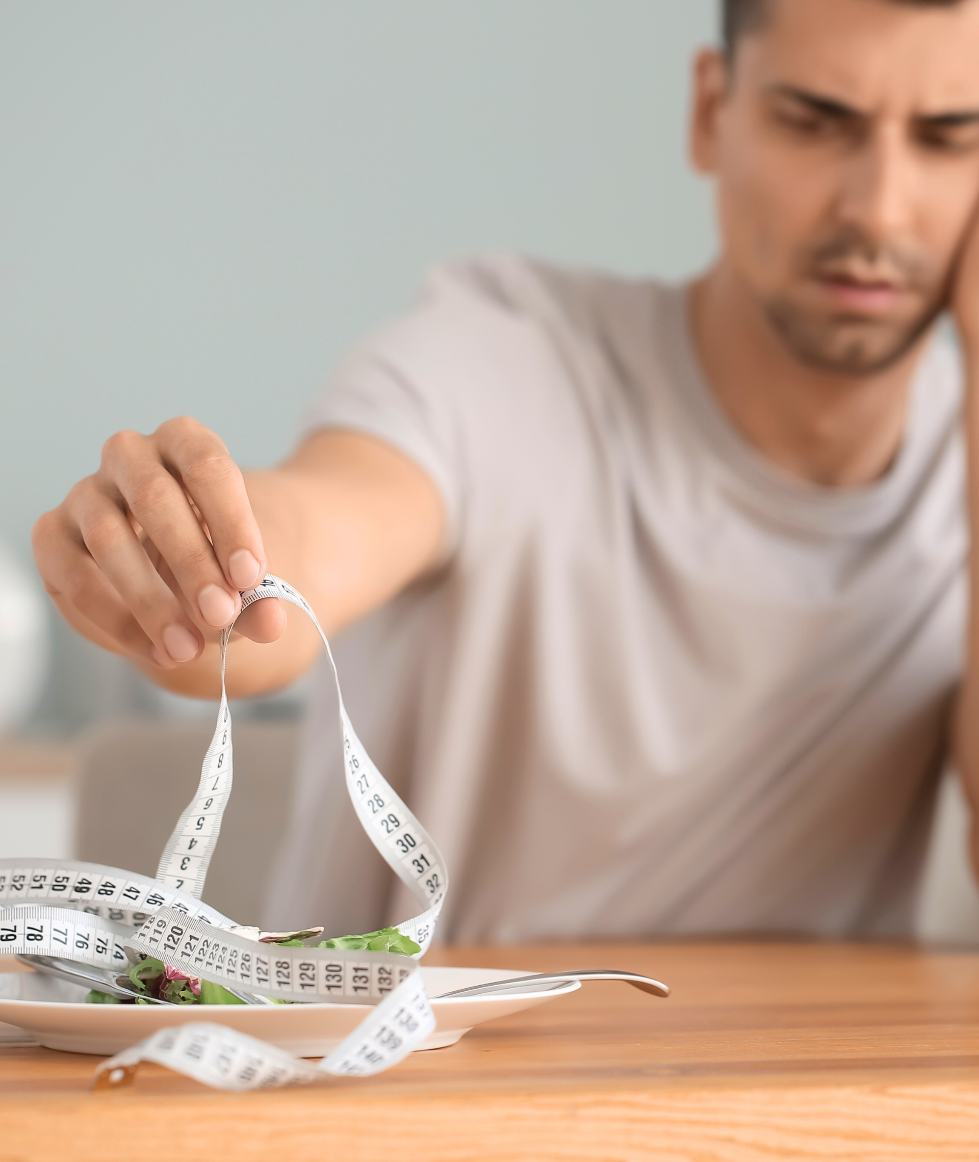 Man looks sadly at a plate of food, tangled with a measuring tape, indoors.