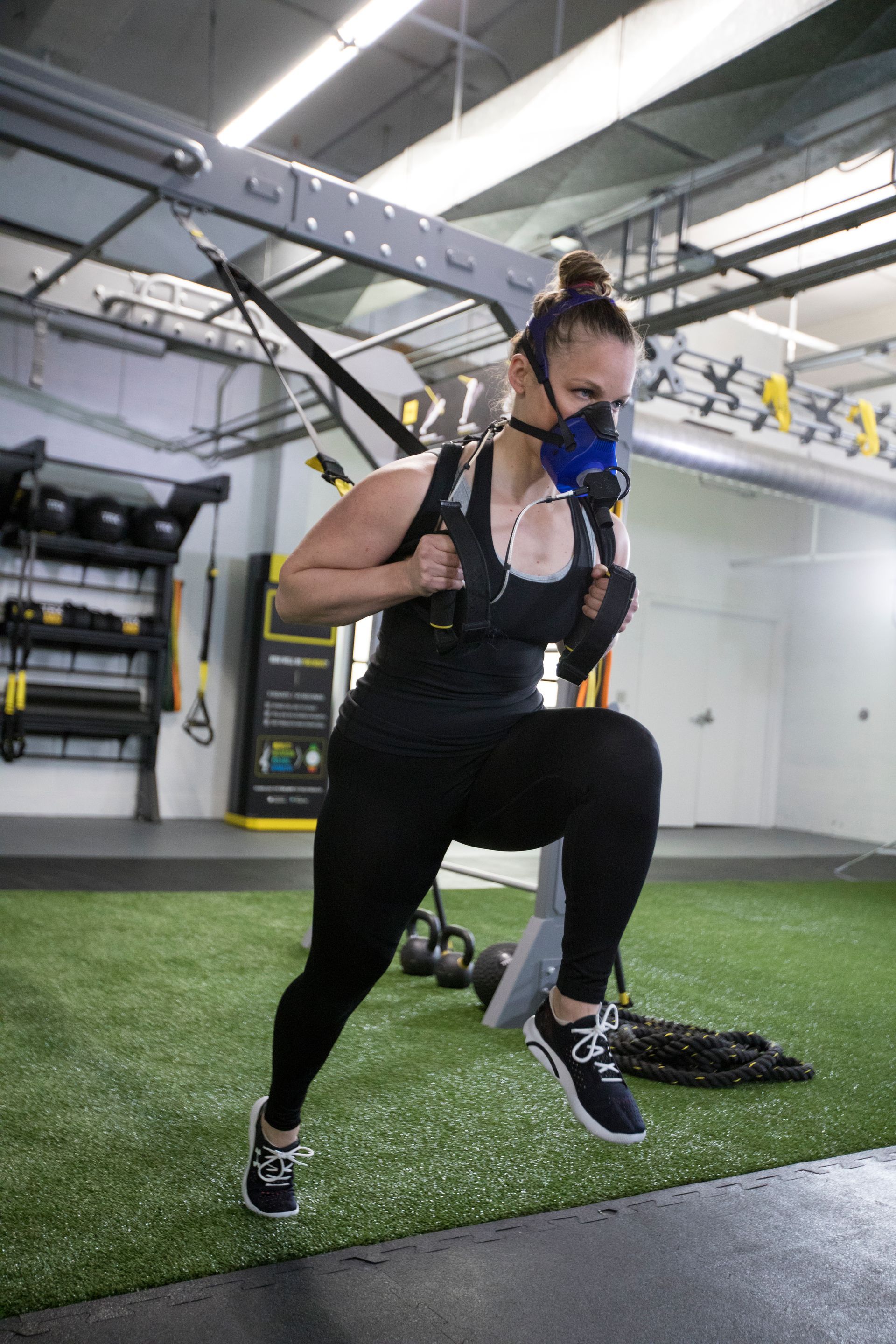 Woman wearing a mask does a TRX exercise in a gym. She is wearing black athletic wear.