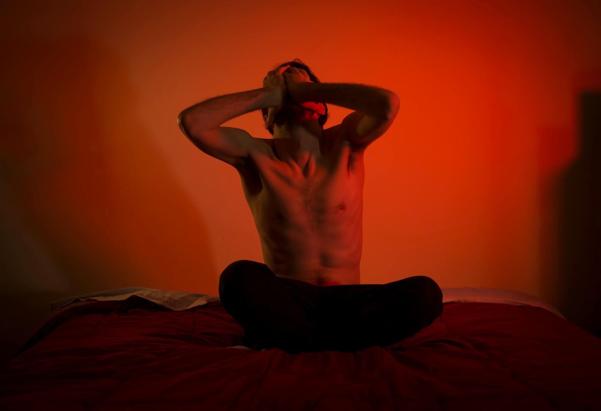 Man sitting cross-legged on a bed, hands on head, lit by orange light, appearing distressed.