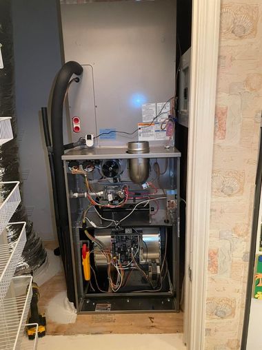 Furnace in a closet, open for inspection. Metal components, wiring visible. Blue light indicates operation.