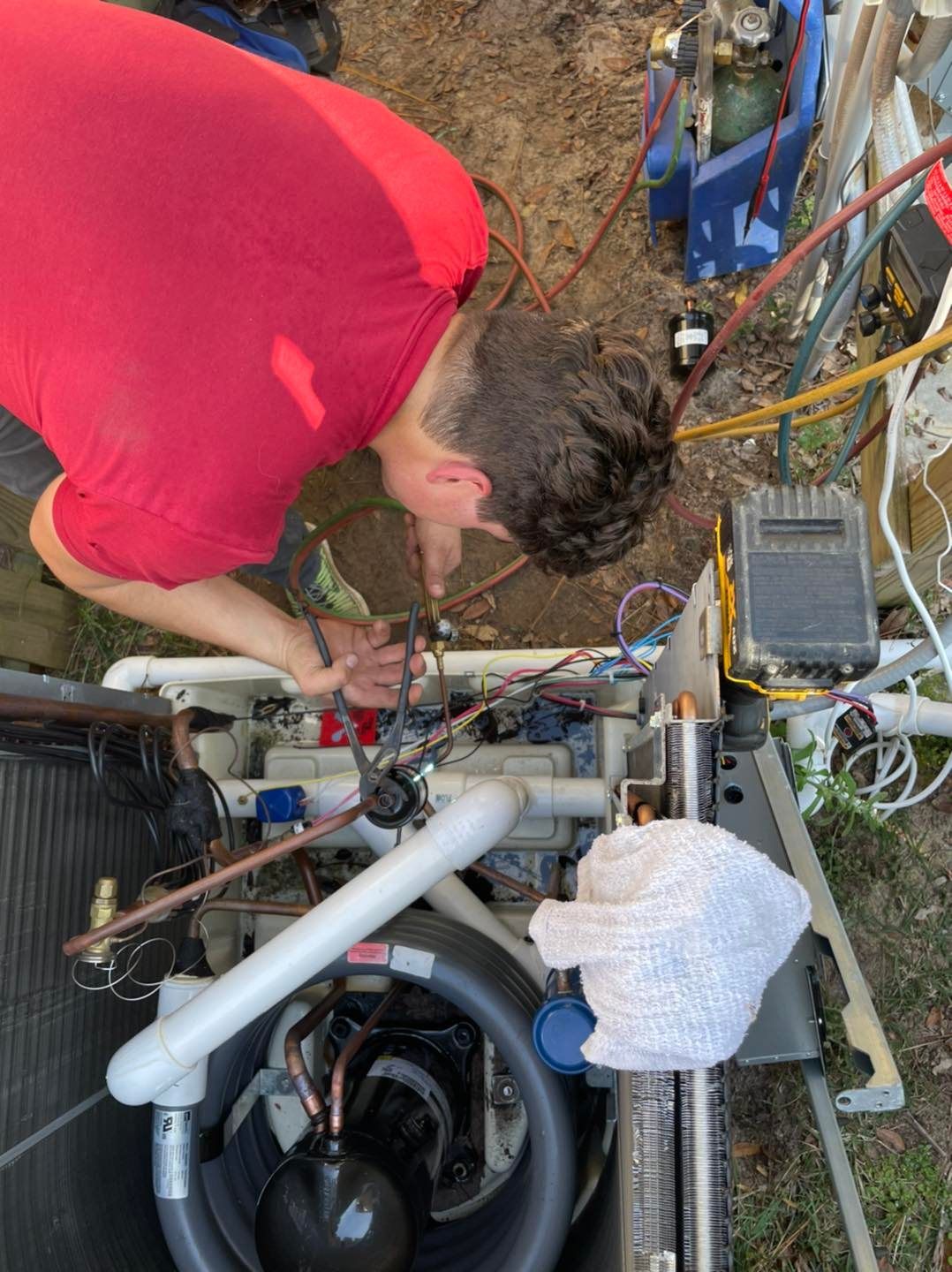 Person in red shirt working on air conditioning unit outdoors.