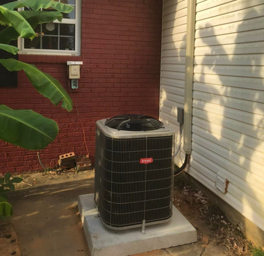 An air conditioning unit outdoors, next to a brick wall and siding.