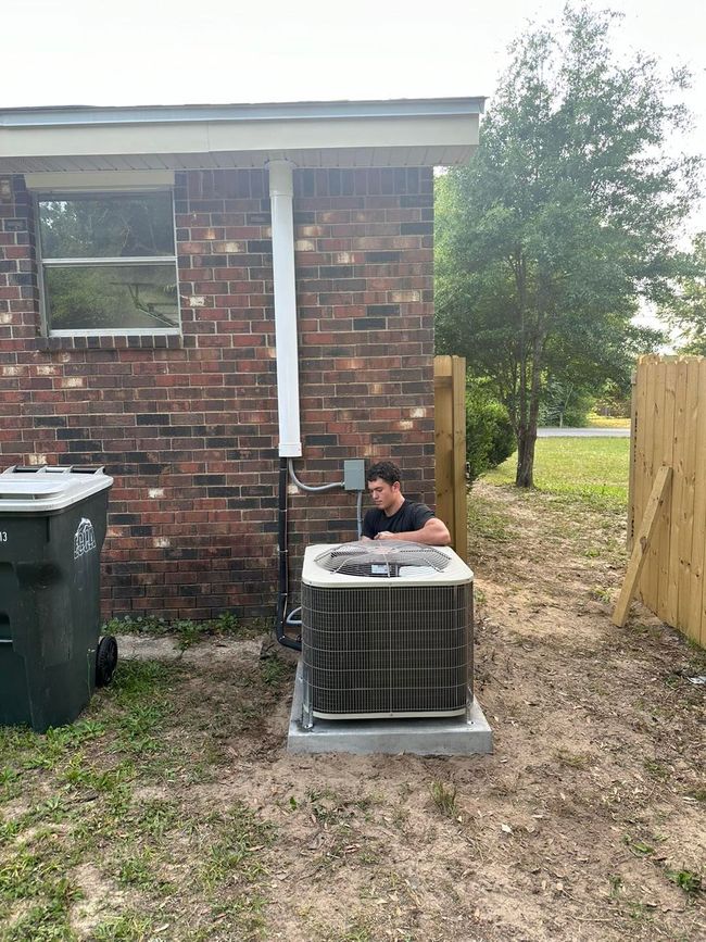 A person works on an air conditioning unit outside a brick building.