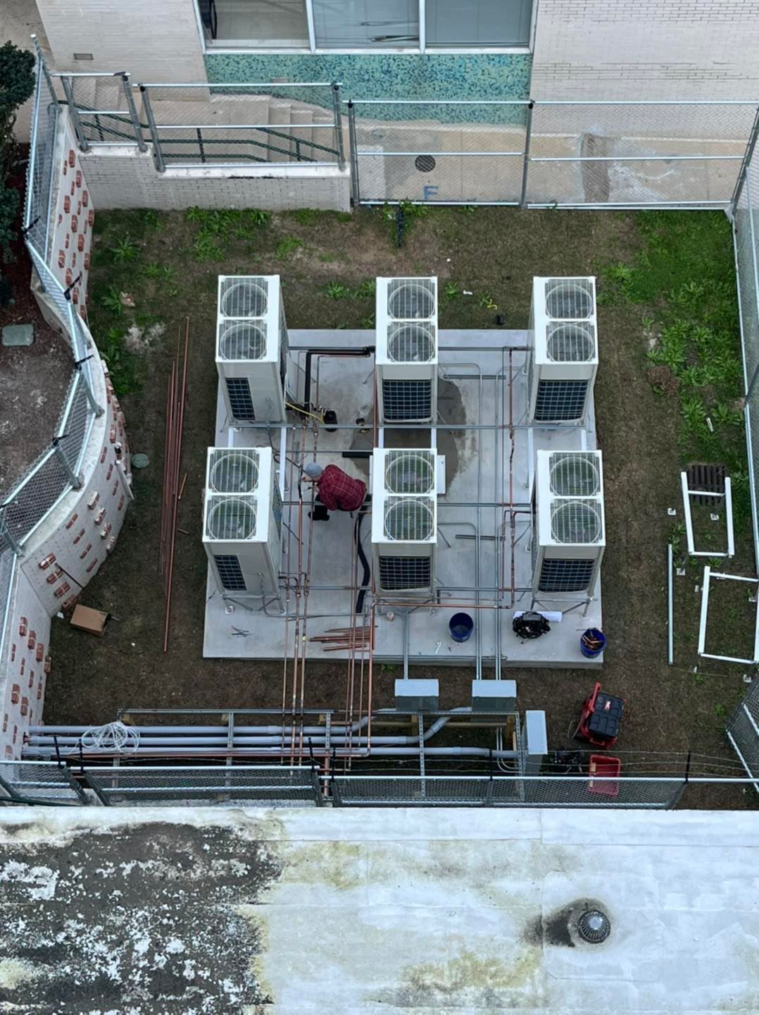 Overhead view of HVAC units on a concrete platform; a person works on the equipment surrounded by grass and fencing.