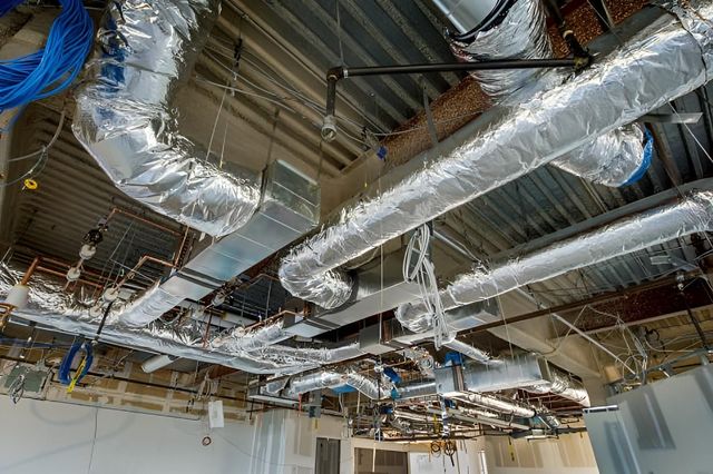 HVAC ductwork and pipes on a ceiling in a construction zone. Silver insulation wraps the ducts.