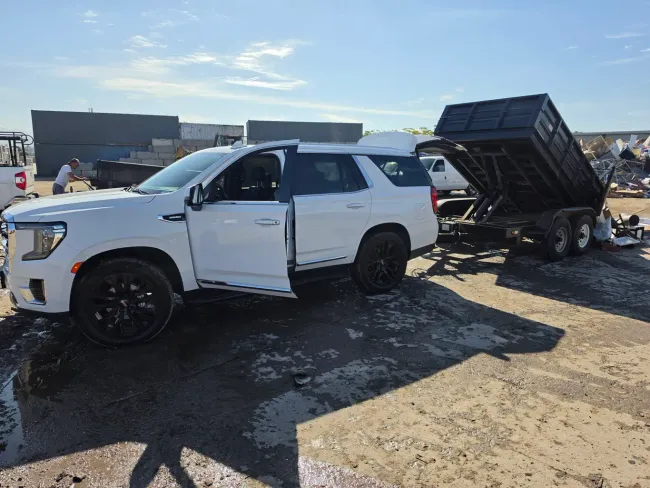 White SUV towing a black dump trailer on a muddy lot under a clear blue sky