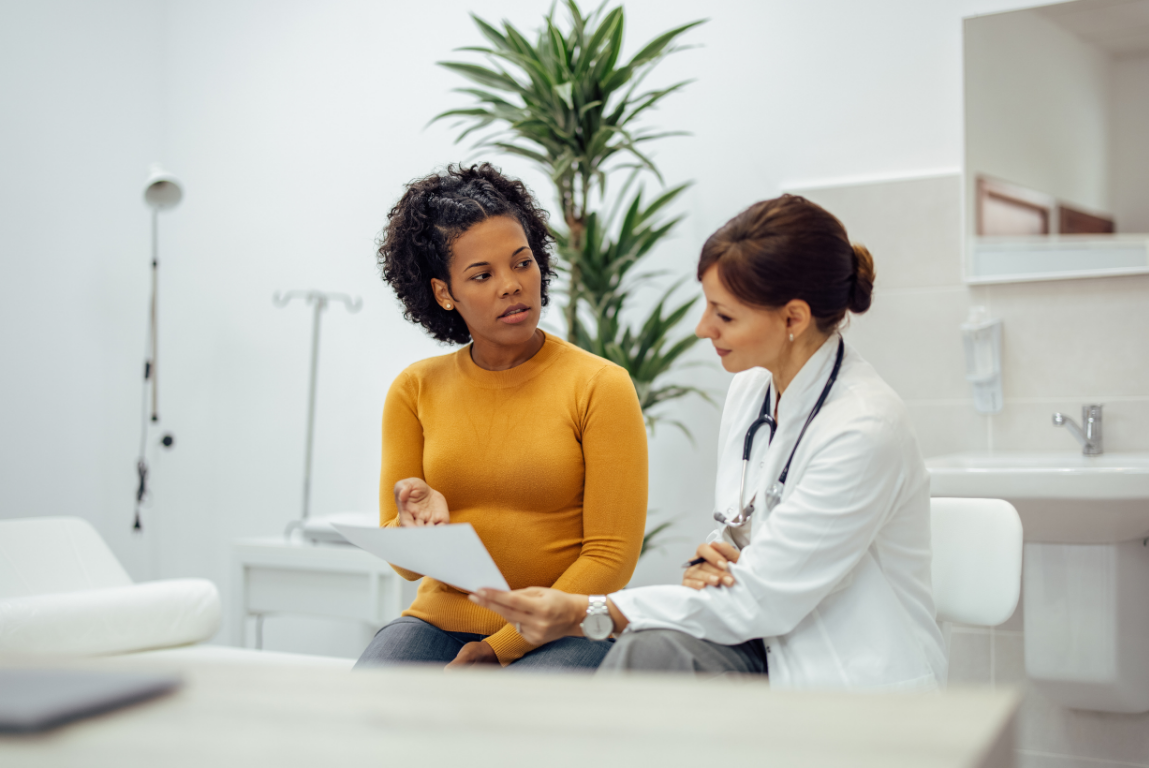 A doctor in a white coat reviews paperwork with a patient in an exam room.