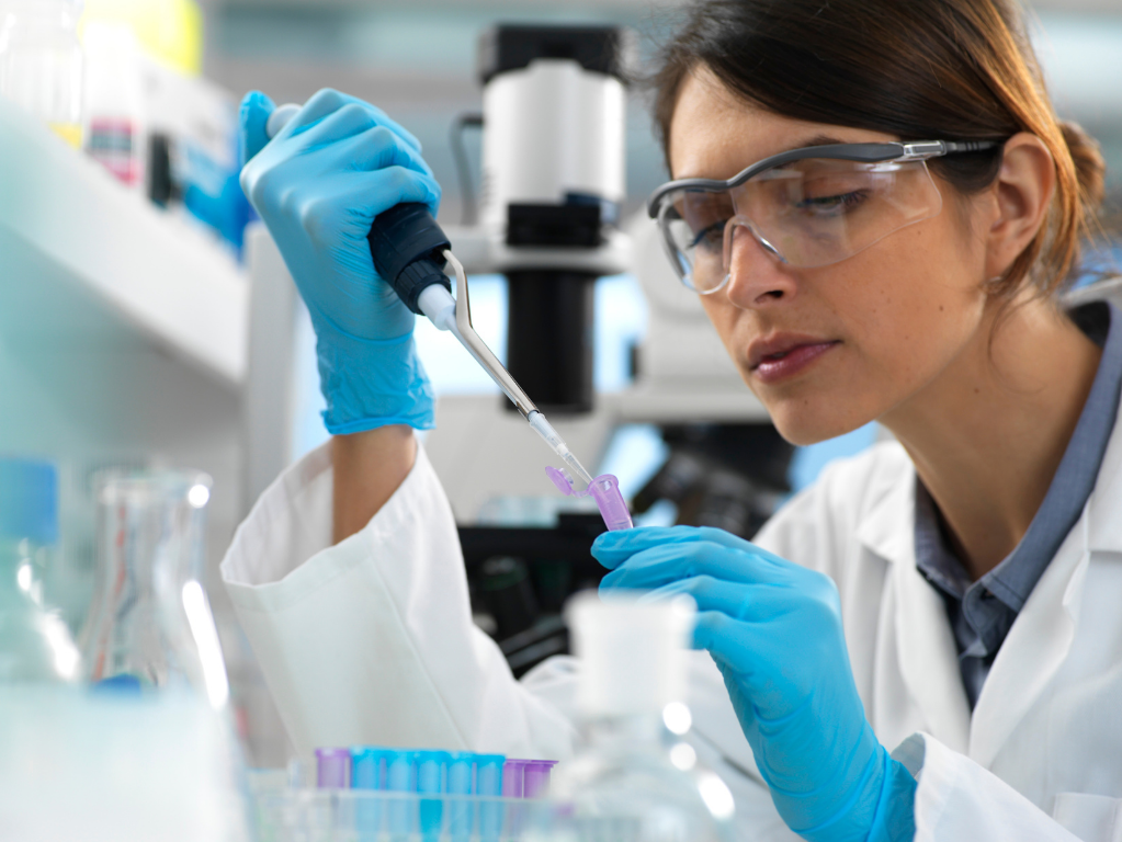 Scientist in lab coat, goggles, and gloves using a pipette to transfer liquid into a test tube.