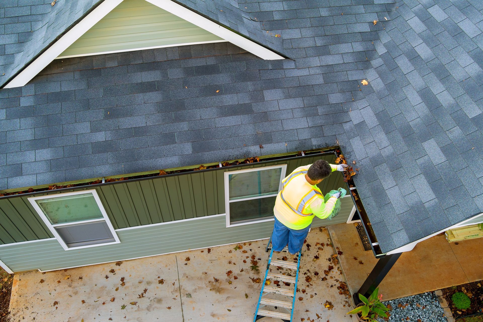 Worker on a roof using a ladder to repair shingles near a gutter.