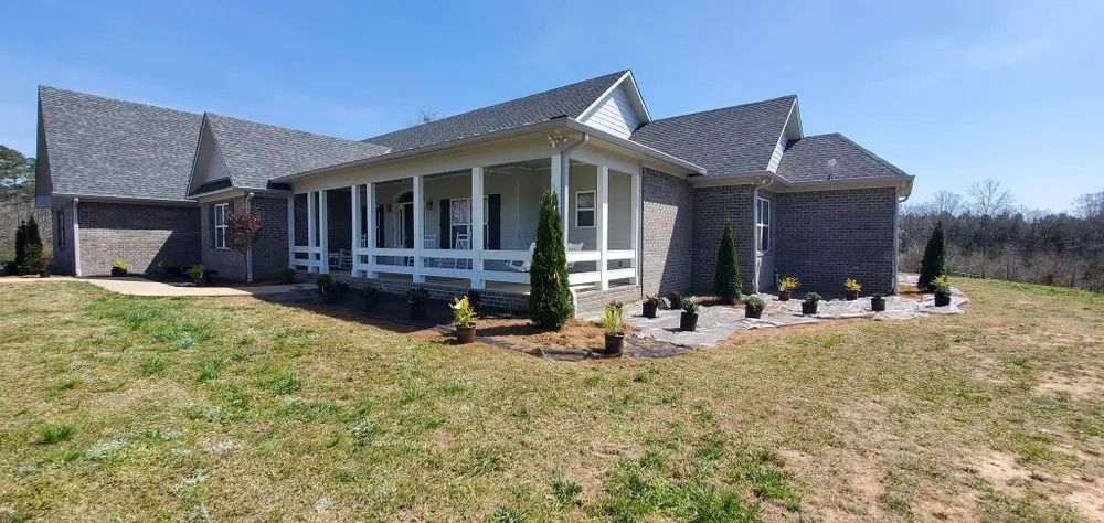 A brick house with a wrap-around porch and dark roof, set on a grassy lot under a blue sky.