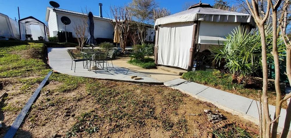 Backyard with concrete paths, gazebo, table, chairs, and small white buildings under a blue sky.
