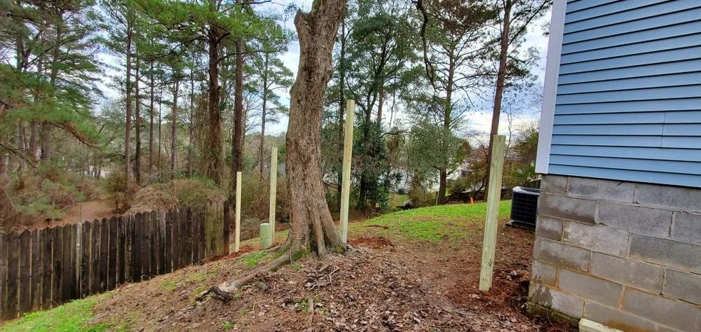 Wooden fence posts installed on a sloped yard near a tree and a blue-sided building.