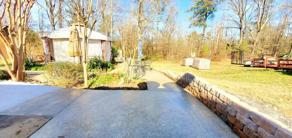 A paved walkway leads to a small building in a sunny, outdoor setting with trees and a stone wall.
