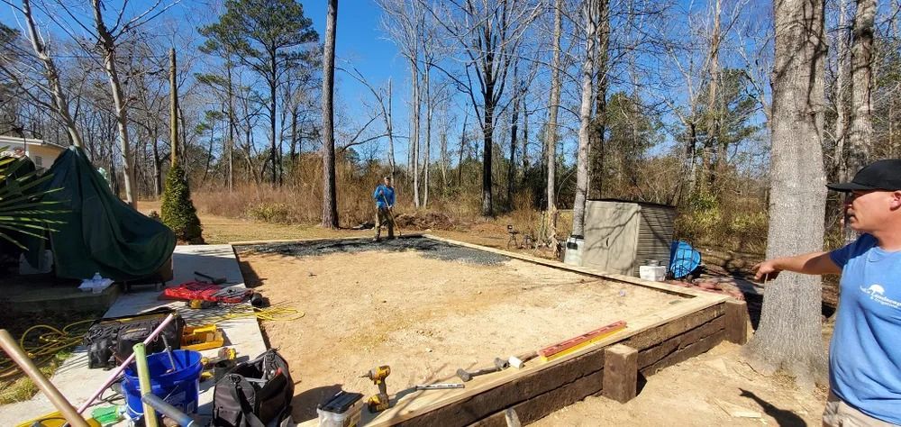 A man points at a sandy area framed by wood. A person stands in the background. Trees fill the scene.
