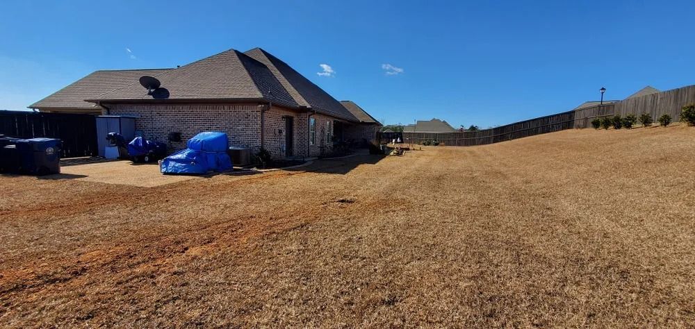 A house with a large, brown yard under a blue sky. A fence and some bushes are in the distance.