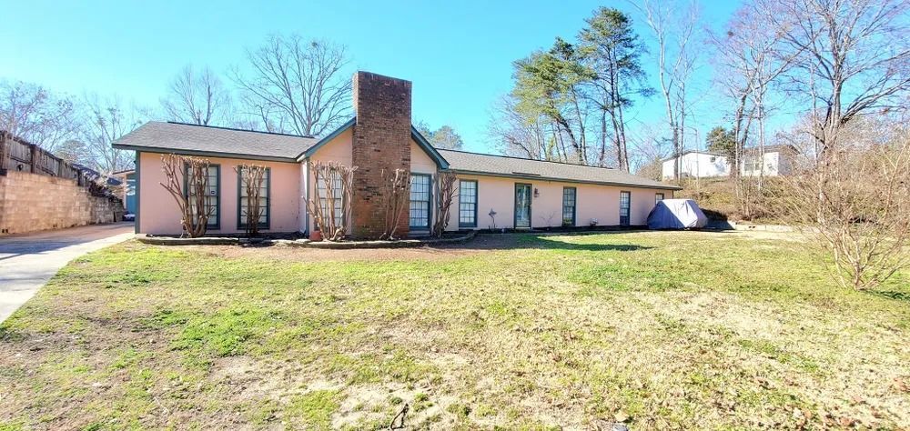 A single-story pink house with a brick chimney sits on a grassy lot. Bare trees and a blue sky are in the background.