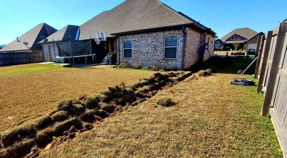 A trench dug in a backyard next to a brick house, with dry grass and a wooden fence.