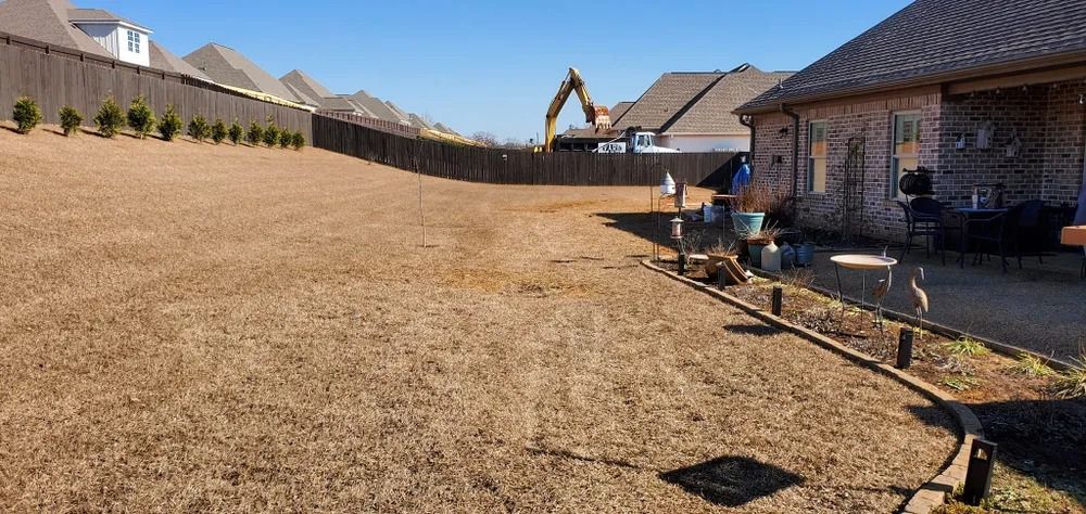 A backyard with brown grass, a wooden fence, and a house. An excavator is in the distance.