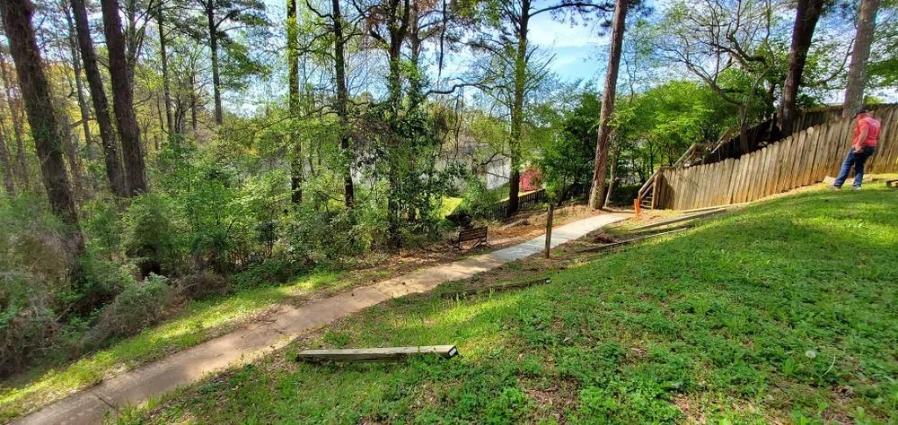 Dirt trail winding through trees and green grass with a person standing near a wooden fence.