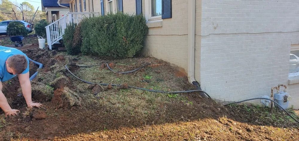 Man working in a dirt garden bed near a house. He's digging. The soil is brown and there's a bush.