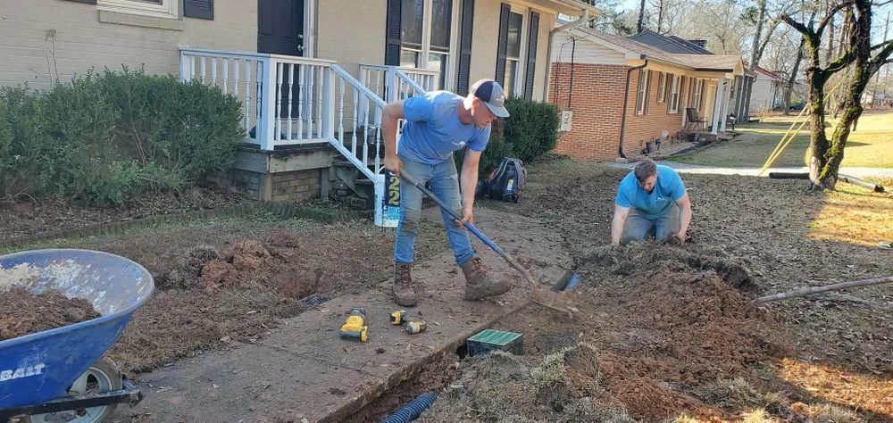 Two men digging in a yard, preparing to lay pipes near a house.