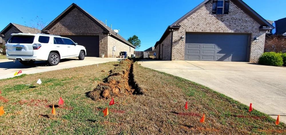 A trench dug in a lawn in front of two houses, marked with flags. A white SUV is parked on the left.