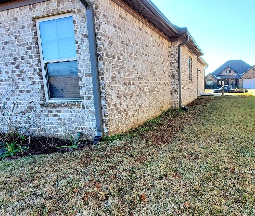 Brick house exterior with window and gutter. Grass yard with bare patches.