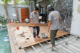 Two workers installing wooden deck planks near a pool, one carries a plank, the other observes.