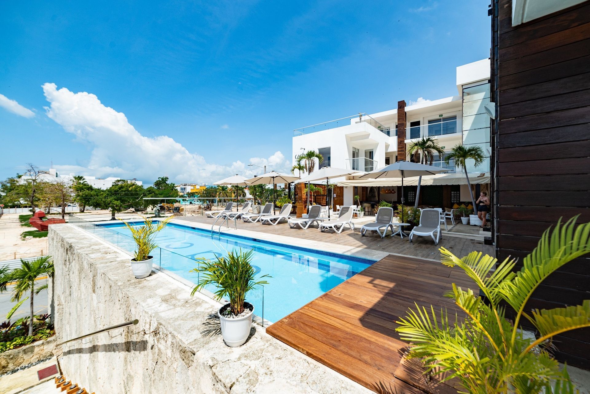 A swimming pool with lounge chairs on a deck, beside a white hotel building under a bright blue sky.