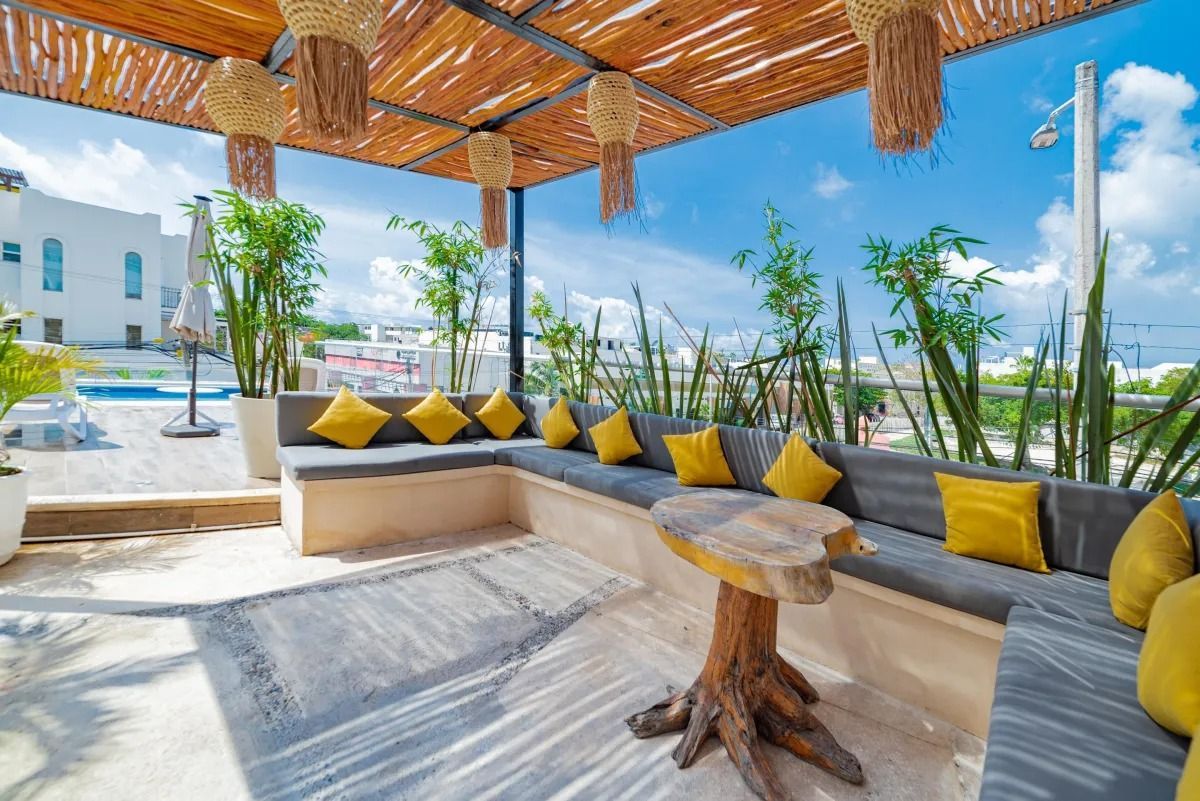 A shaded outdoor lounge with stone benches, gray cushions, yellow pillows, a rustic wooden table, and potted cacti.